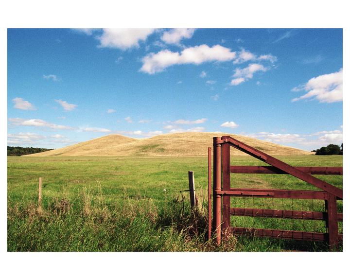 A red wooden gate stands in a green field with rolling hills under a blue sky with white clouds.