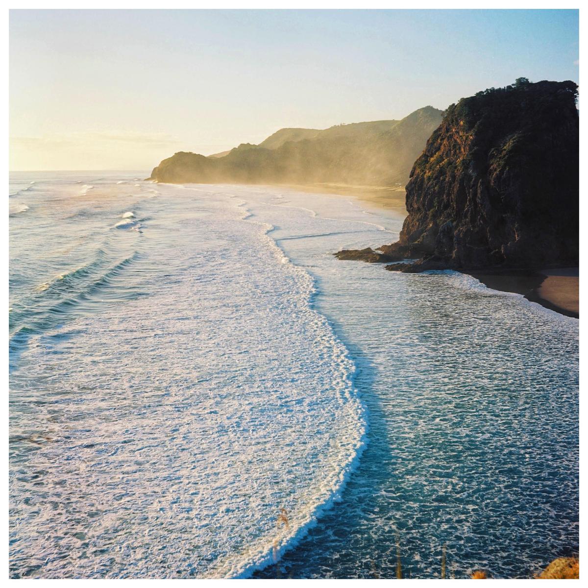 Waves crash on a beach next to a large rock formation and hills in the distance.