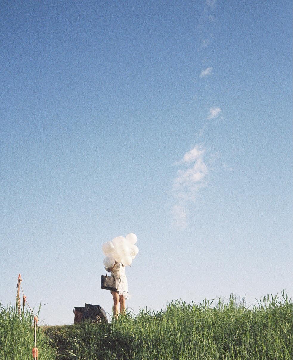 A woman stands in a grassy field holding a cluster of white balloons and a handbag under a clear blue sky.