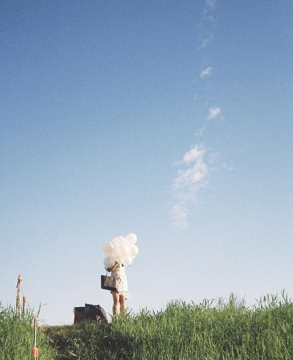 A woman stands in a grassy field holding a cluster of white balloons and a handbag under a clear blue sky.