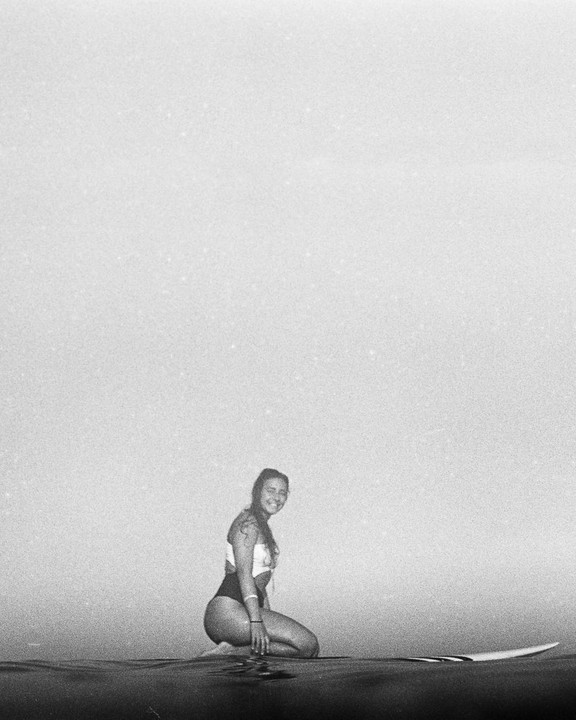 A woman kneels on a surfboard in the water, smiling at the viewer.