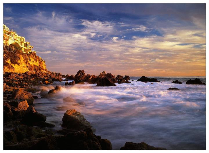 A building sits atop a cliff overlooking a rocky coastline with waves crashing against the shore under a dramatic sky.