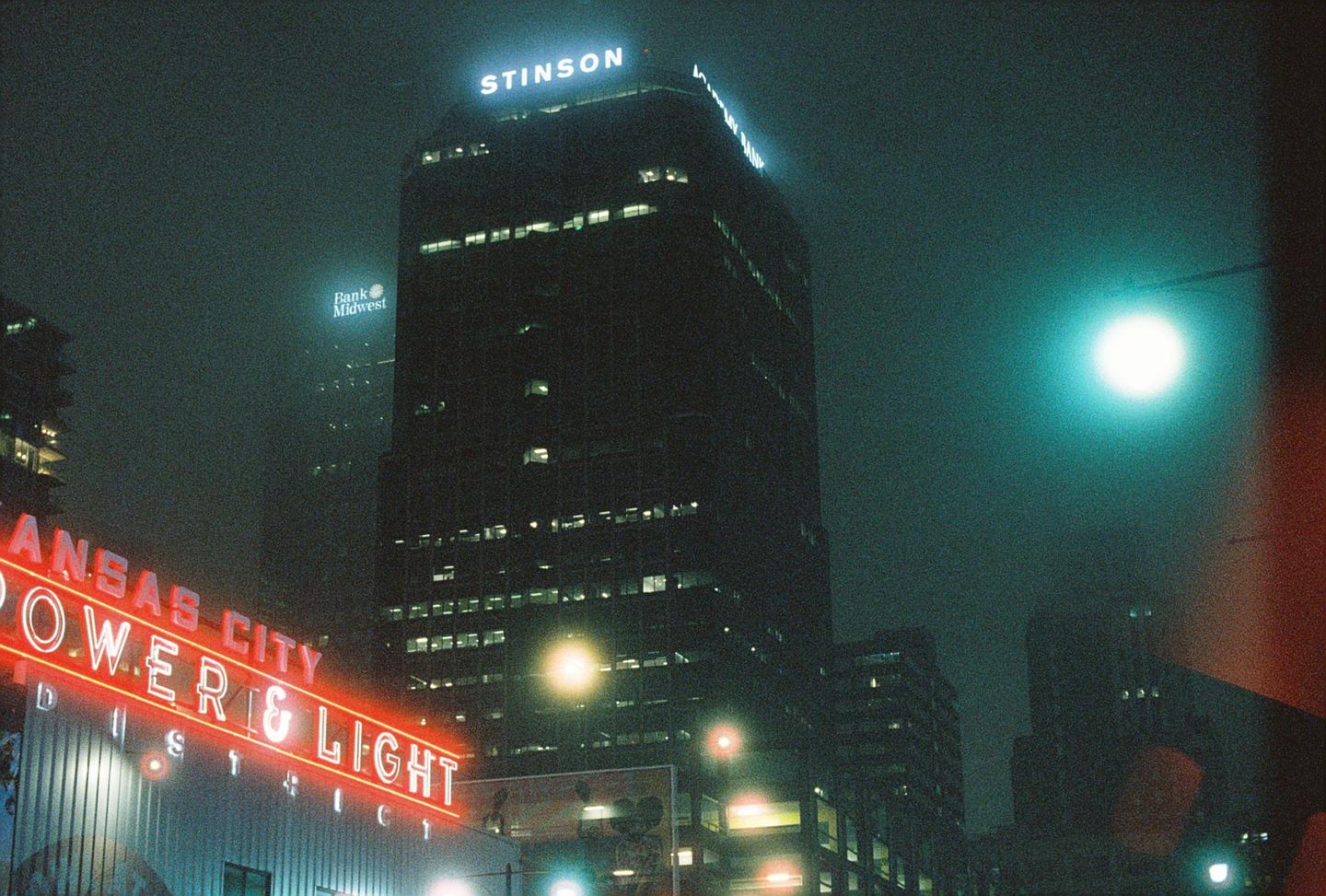 A tall building with a glowing sign for Stinson stands against a foggy night sky, with a red neon sign for Kansas City Power & Light in the foreground.