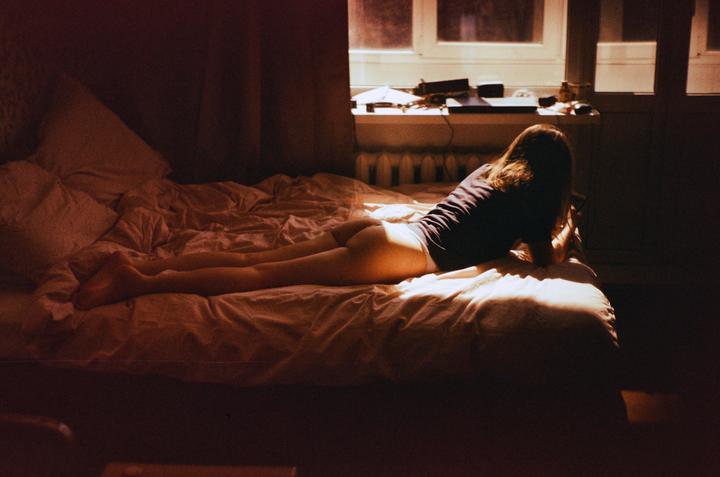 A woman lies on a bed in a dimly lit room, illuminated by a shaft of sunlight from a window.