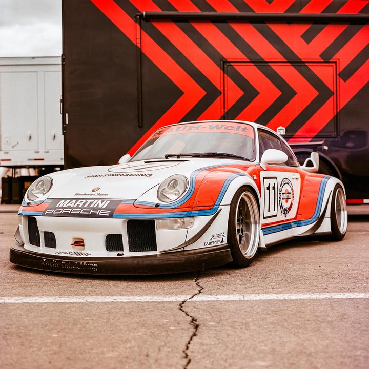 A white Porsche race car with red and blue stripes is parked on asphalt in front of a black and red striped trailer.
