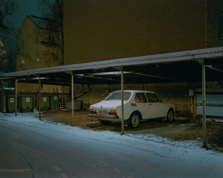 A white car is parked under a carport next to a building on a snowy night.