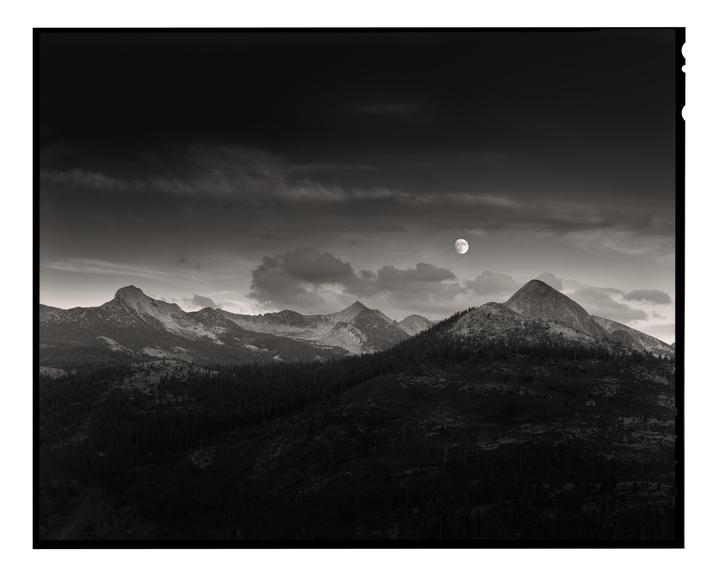 A full moon hangs in a cloudy sky above a mountain range covered in trees.