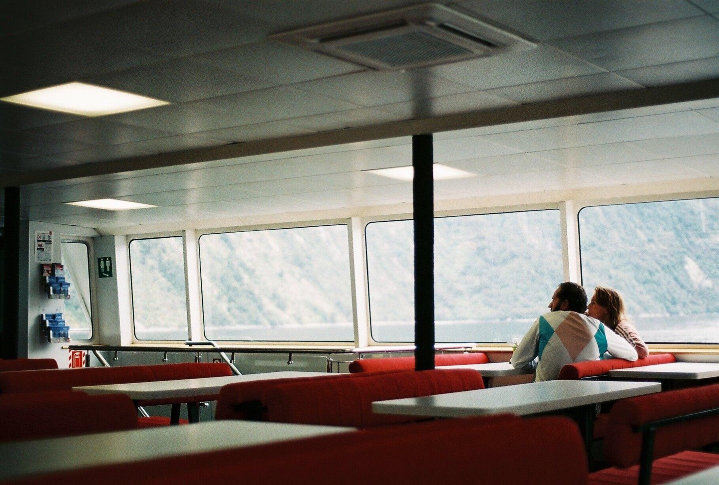 A couple sits at a table on a ferry looking out at a mountain landscape.