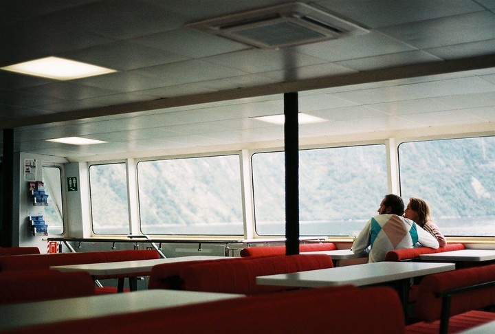 A couple sits at a table on a ferry looking out at a mountain landscape.