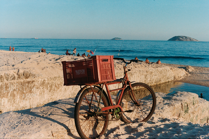 A red bicycle with two crates on its rack is parked on a sandy beach next to the ocean, with people relaxing in the background.
