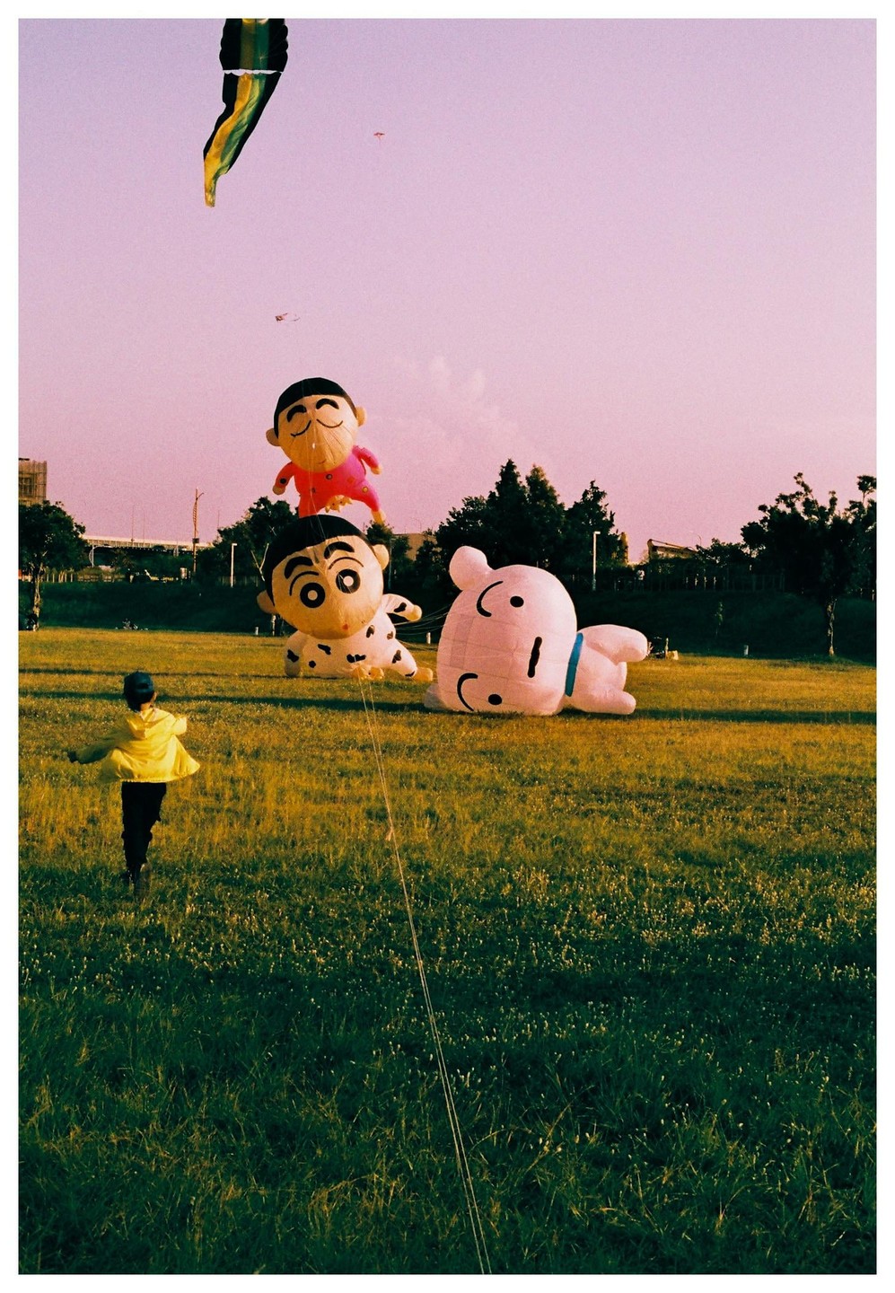 A child in a yellow jacket runs across a grassy field with large cartoon character kites.