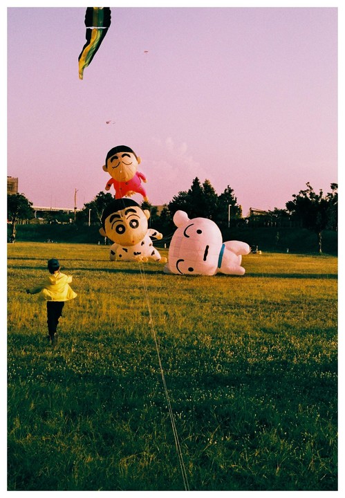 A child in a yellow jacket runs across a grassy field with large cartoon character kites.