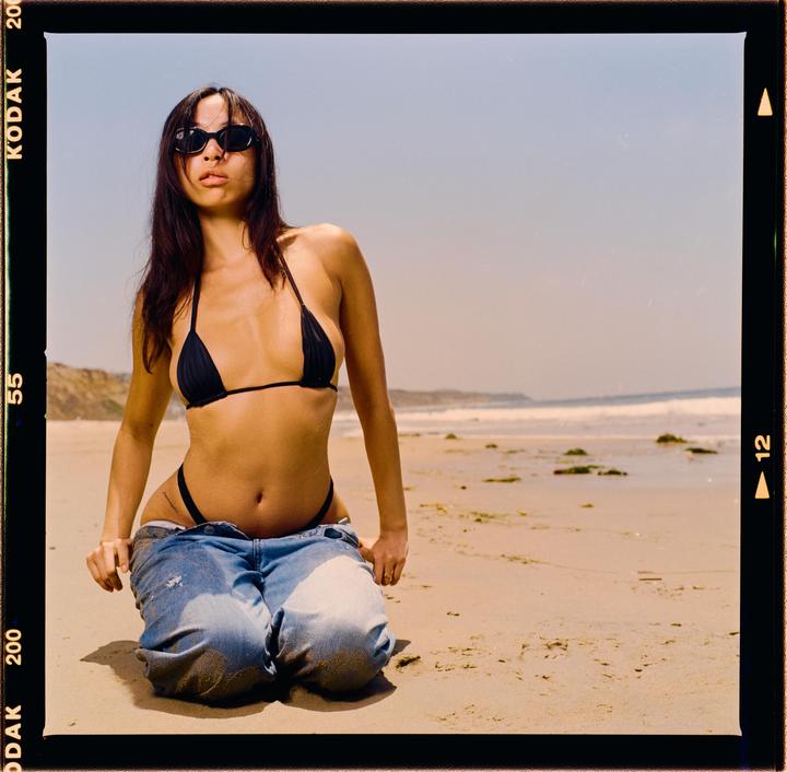 A woman kneels on a sandy beach with her jeans pulled down, wearing a bikini top and sunglasses.
