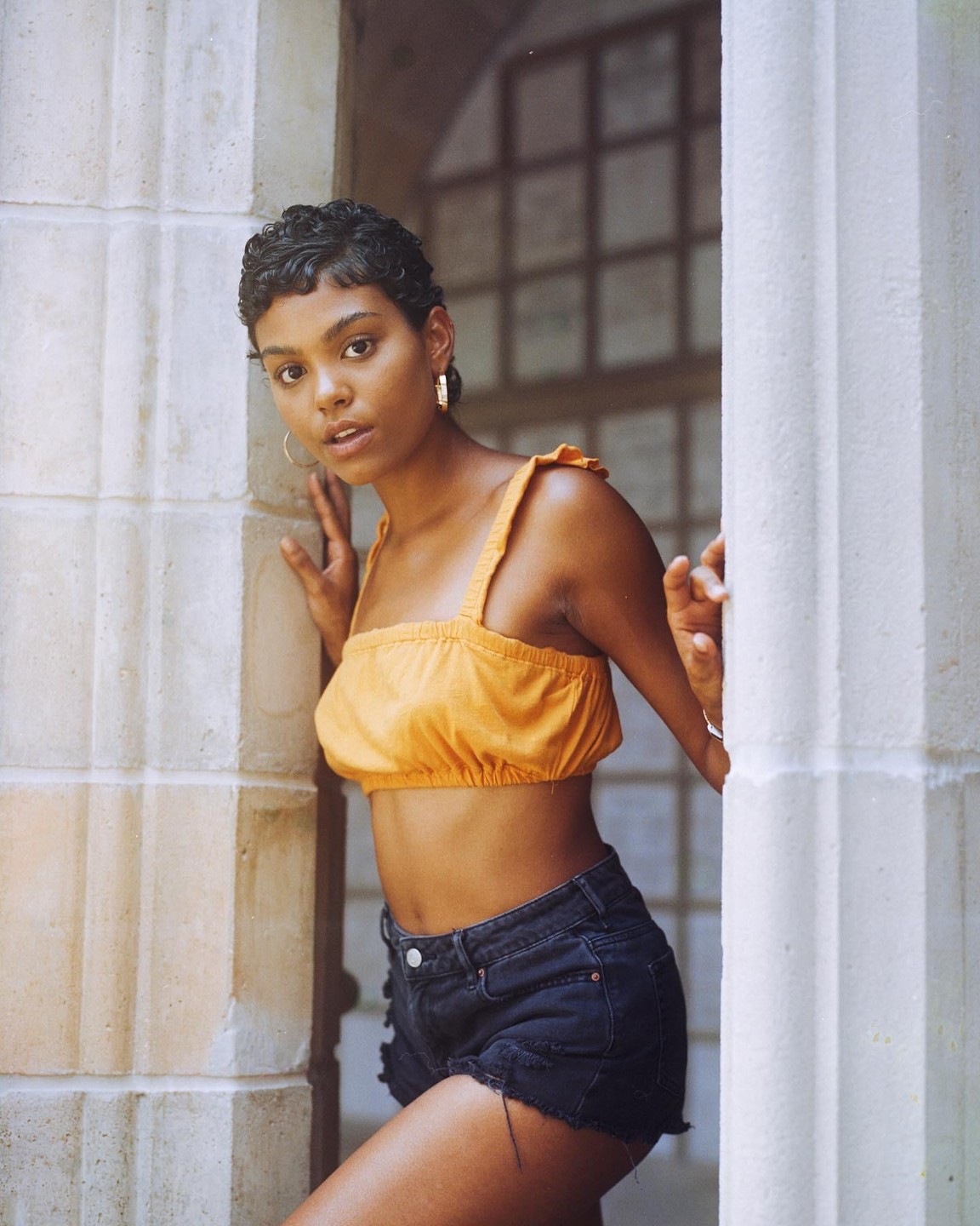 A woman in a yellow crop top and denim shorts leans against a stone wall.