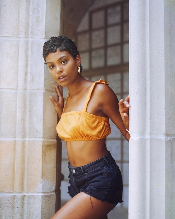 A woman in a yellow crop top and denim shorts leans against a stone wall.