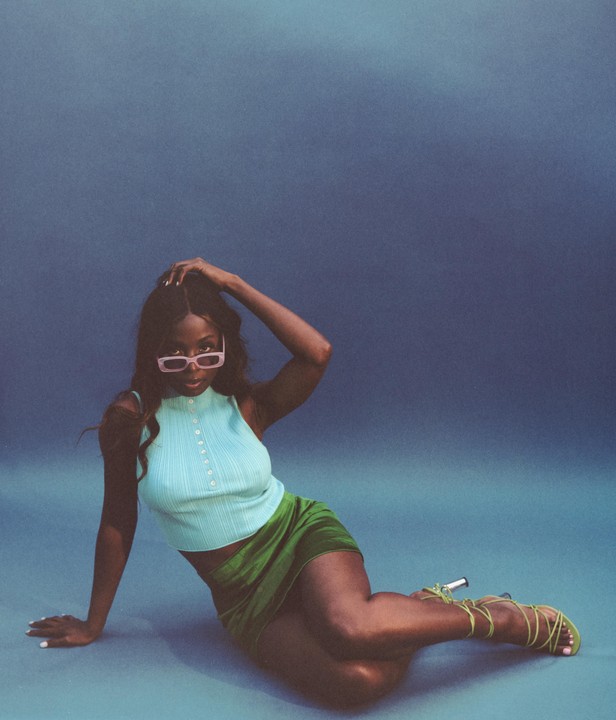 A woman wearing a light blue top, green skirt, and pink sunglasses is posing on a blue surface.