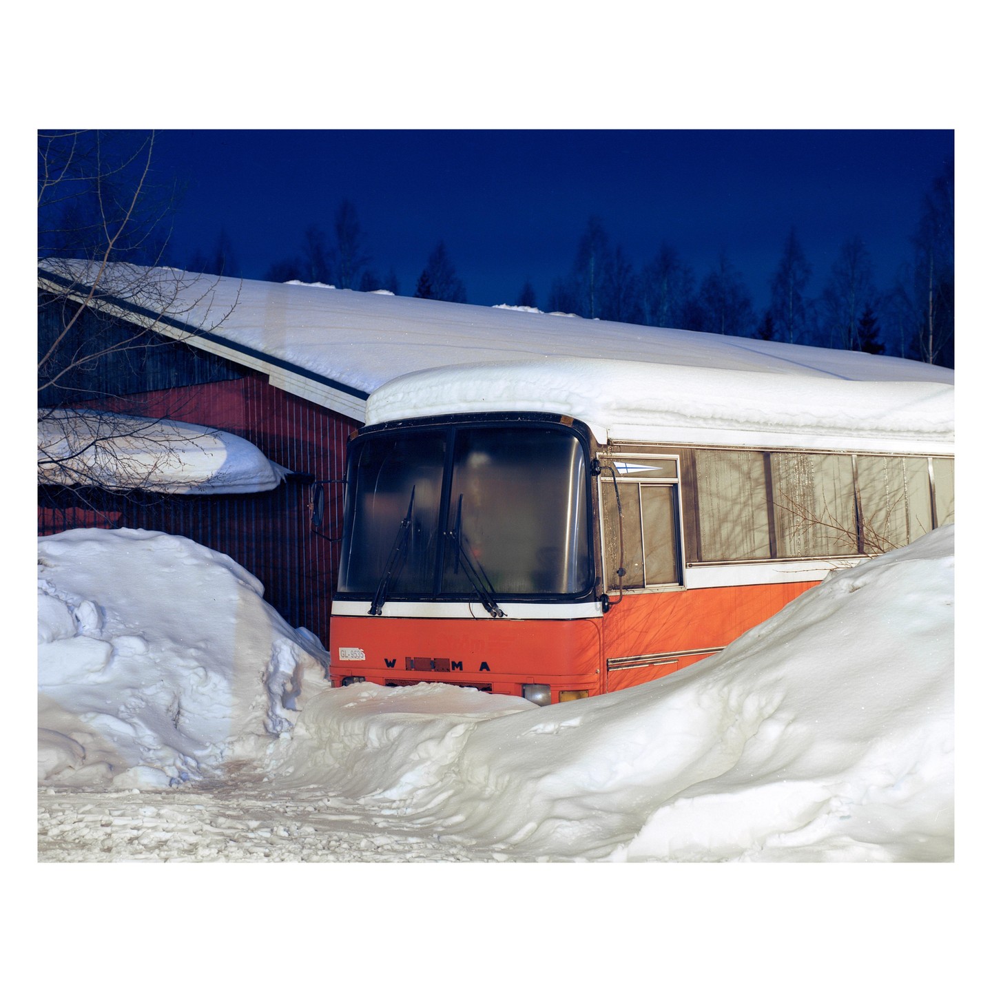 An orange bus is parked next to a red building, surrounded by deep snow under a dark blue sky.