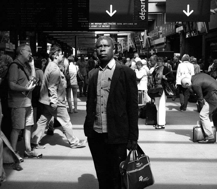 A man stands in a busy train station with a bag in his hand, surrounded by other travelers.