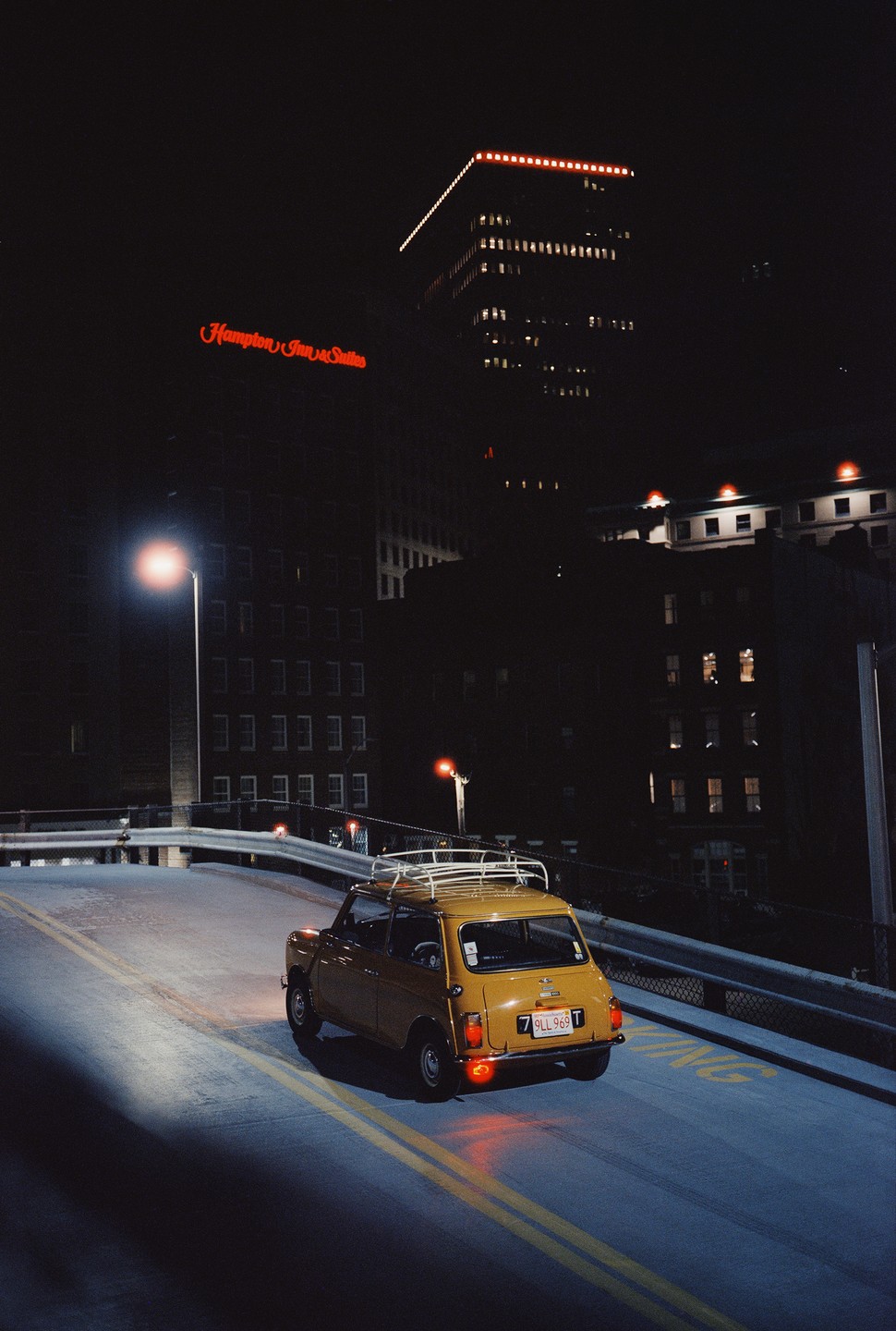 A yellow mini cooper is parked on a ramp at night with a Hampton Inn & Suites building in the background.