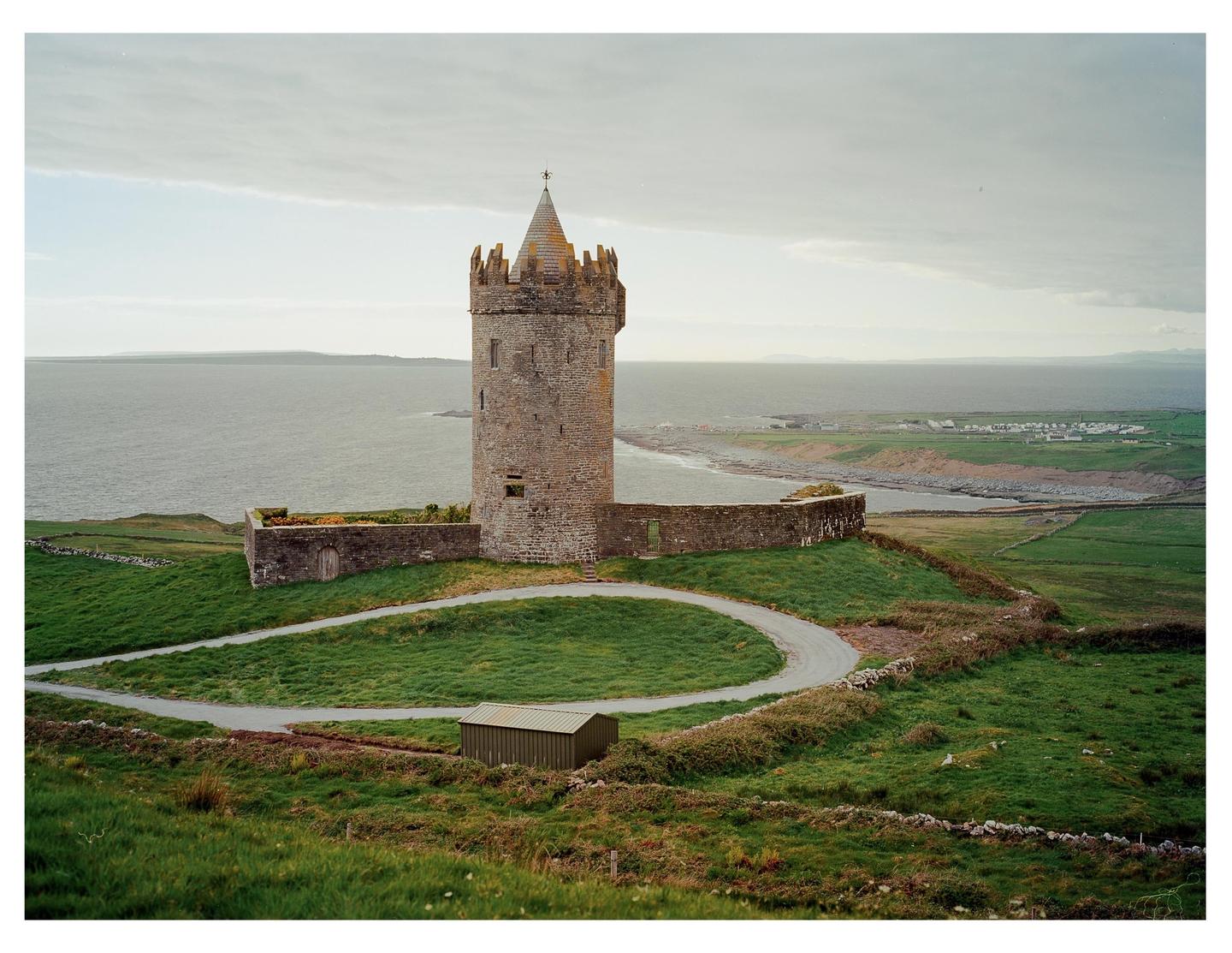 A stone tower with a conical roof stands on a grassy hill overlooking the ocean and a small coastal village.