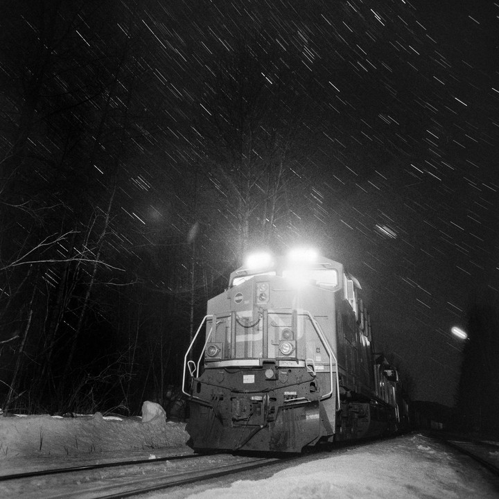 A train with bright headlights sits on snowy tracks at night under a sky filled with star trails.