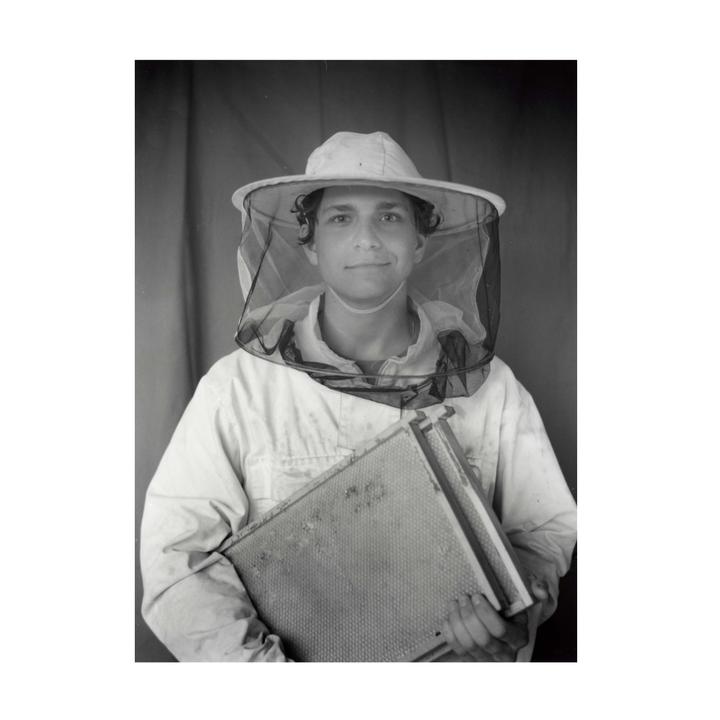 A young man wearing a beekeeper suit and hat holds a honeycomb frame.