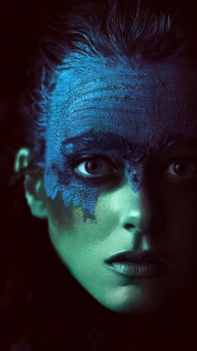 A woman's face is illuminated with blue and green light, with cracked blue paint on her forehead and around one eye.