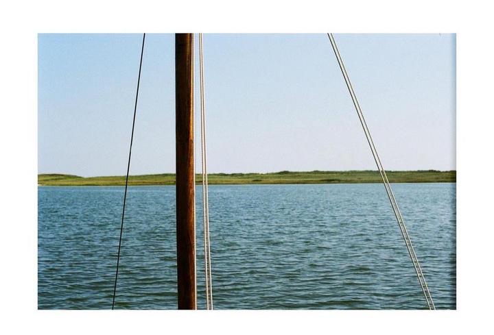 A wooden mast and ropes are visible in the foreground of a body of water with a grassy bank in the distance.