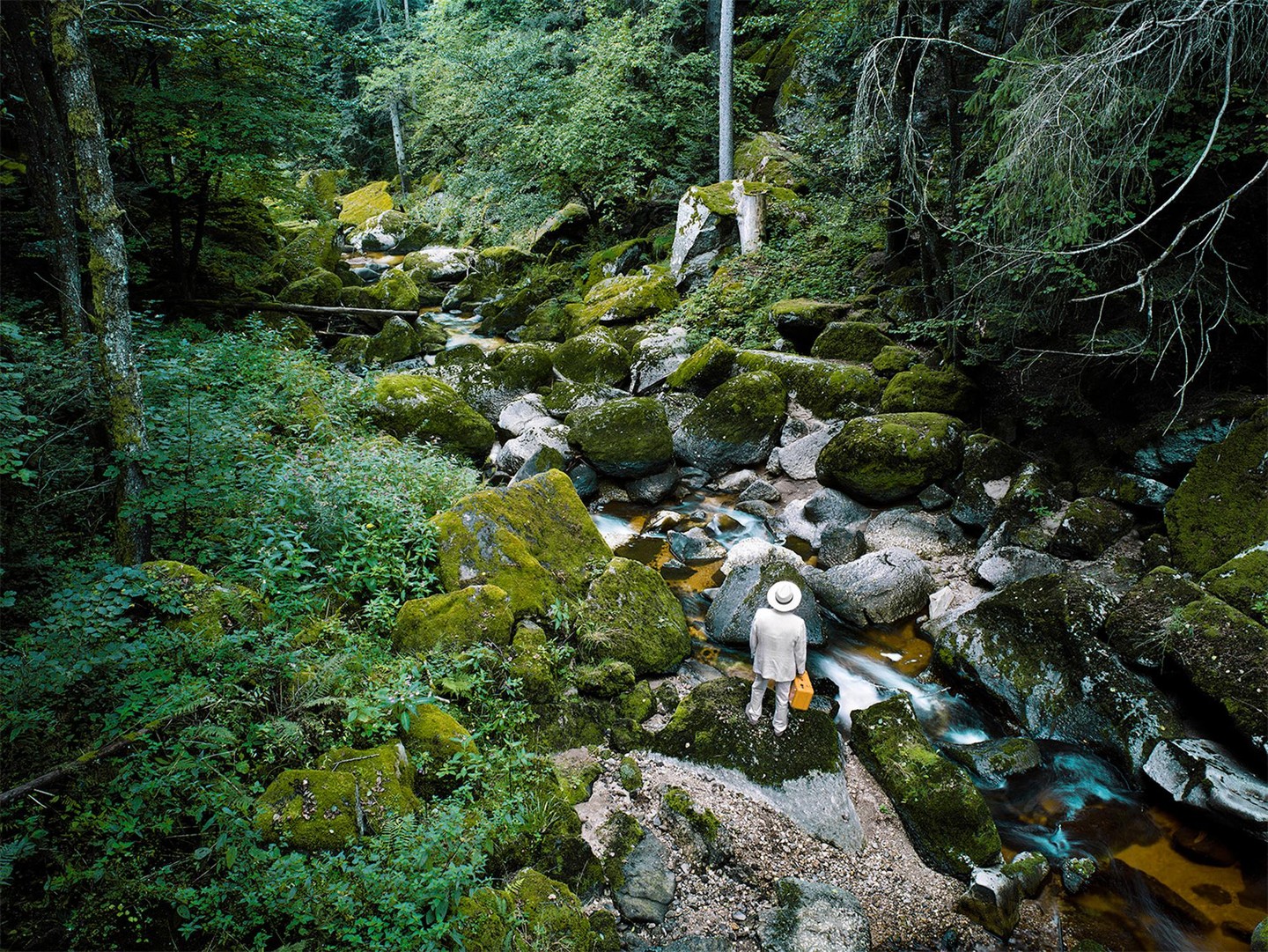 A person in a white suit and hat stands by a stream in a forest with moss-covered rocks.