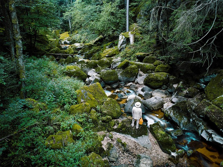 A person in a white suit and hat stands by a stream in a forest with moss-covered rocks.