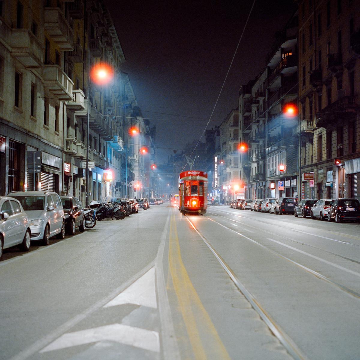 A red tram travels down a street lined with parked cars and buildings at night.
