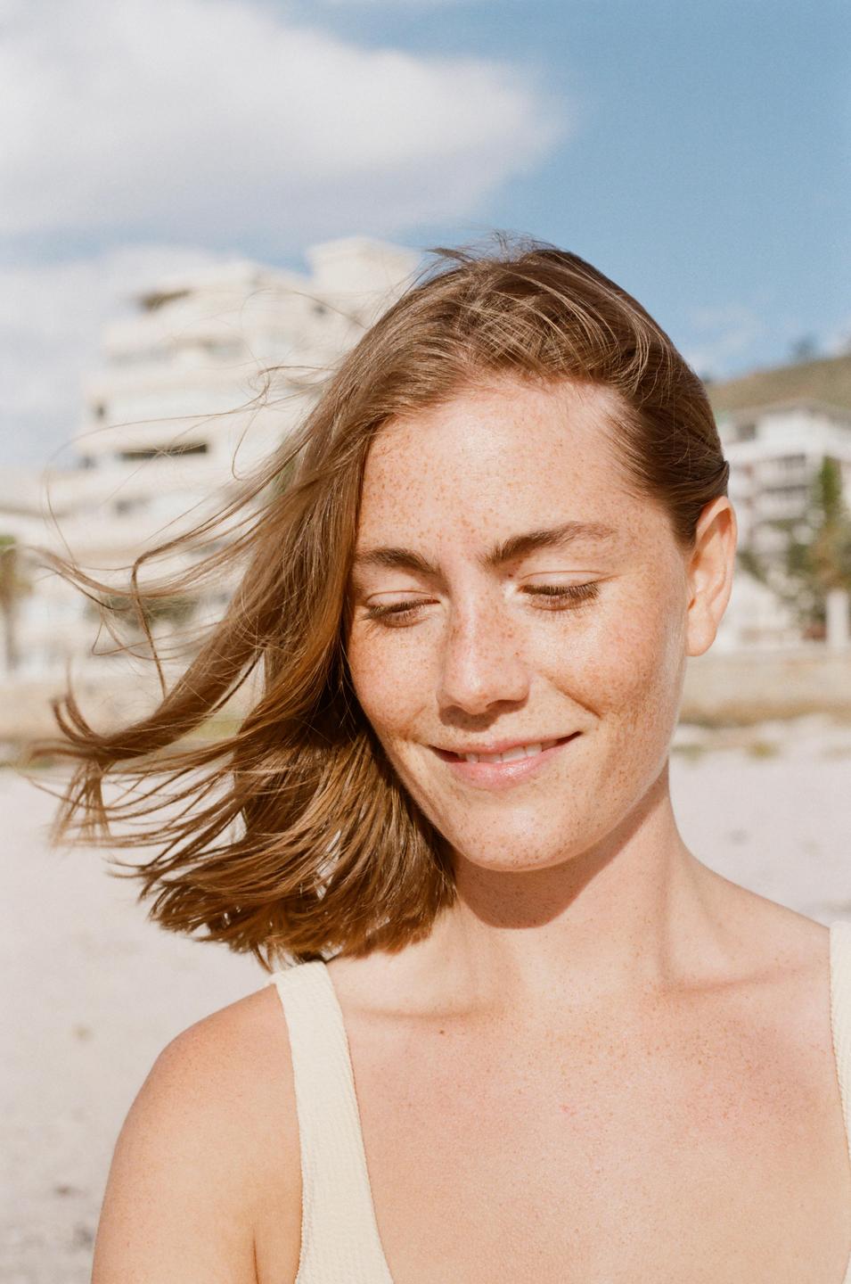 A woman with freckles and closed eyes smiles with her hair blowing in the wind on a beach.
