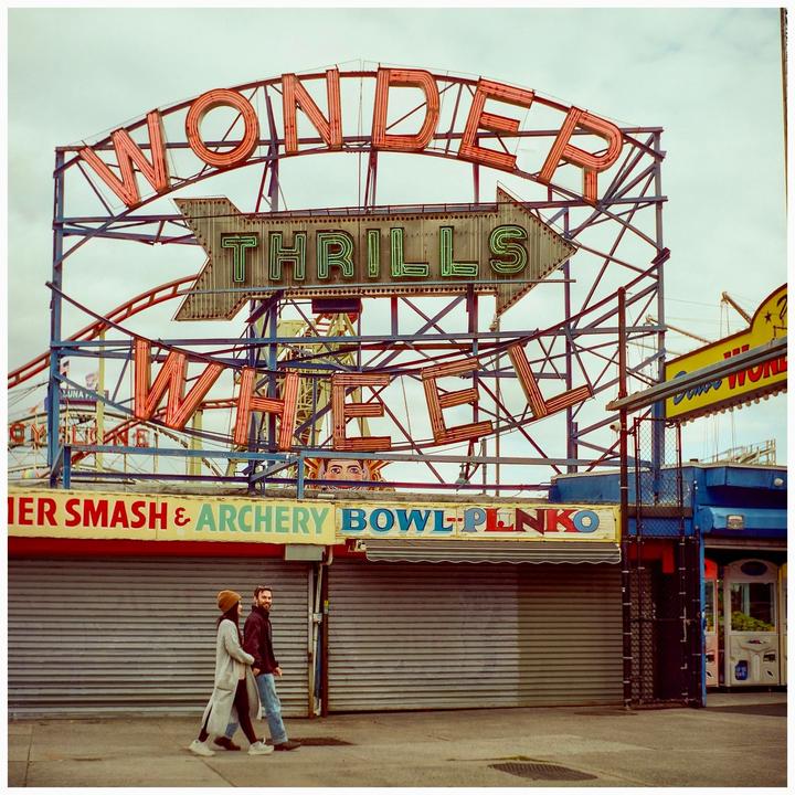 A couple walks past a large neon sign for "Wonder Wheel Thrills" at an amusement park.
