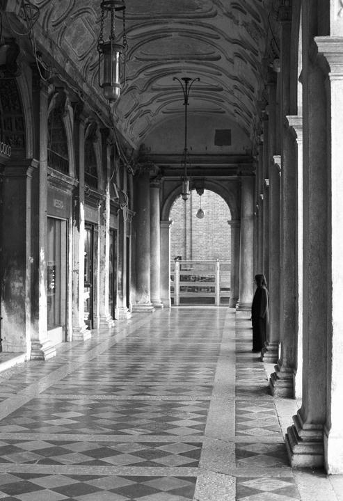 A person stands in a long, arched hallway with a checkered floor and columns.