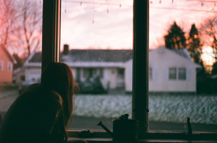 A girl looks out a window at a sunset over a snowy neighborhood.