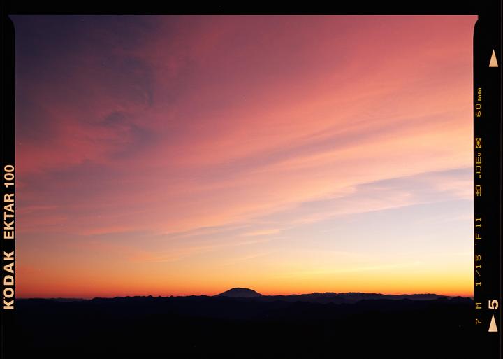 A vibrant sunset sky with hues of pink, orange, and purple stretches above a dark mountain range, with a prominent peak in the center.