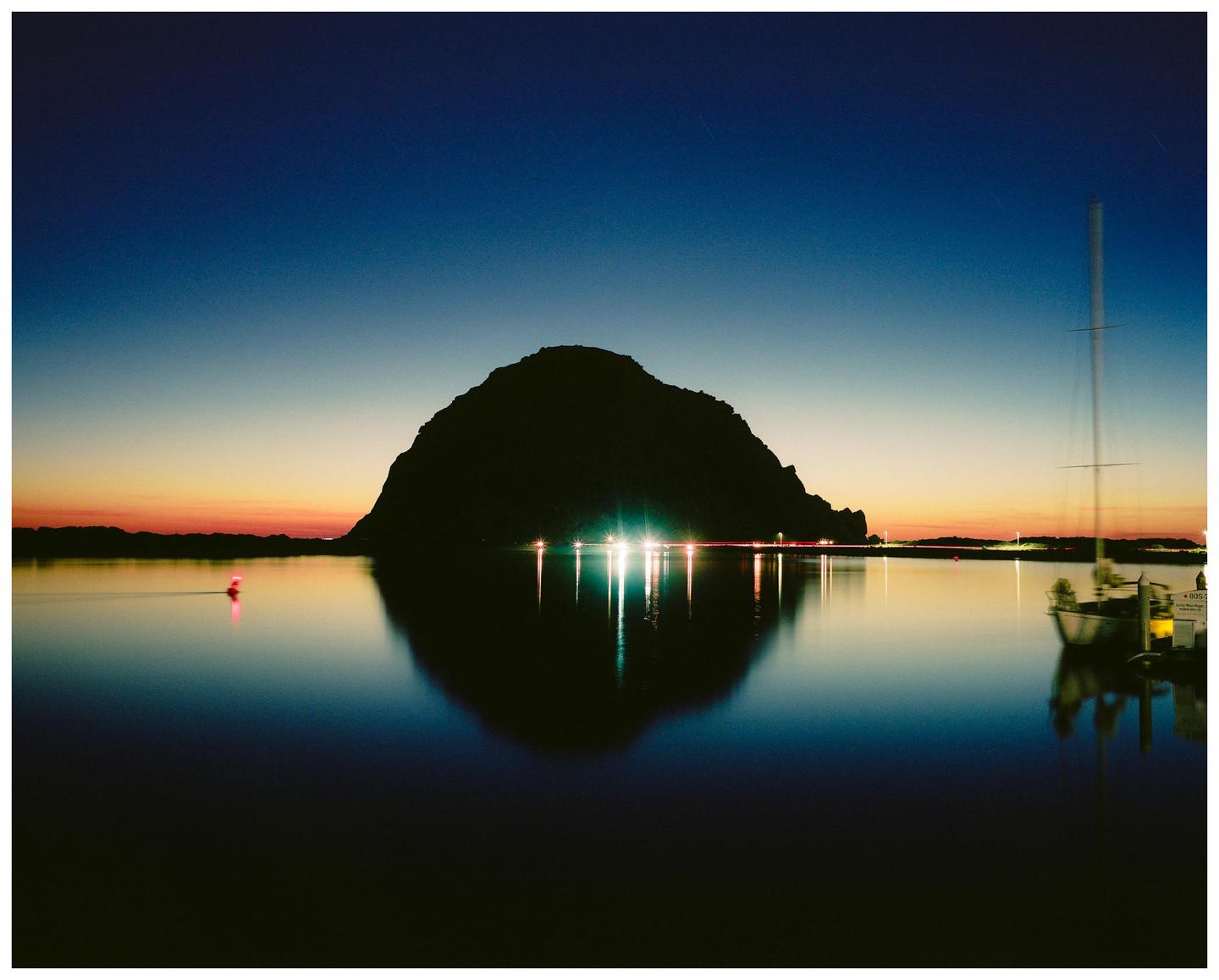 A large rock formation is silhouetted against a colorful sky over water with lights reflecting on the surface and a boat docked on the right.