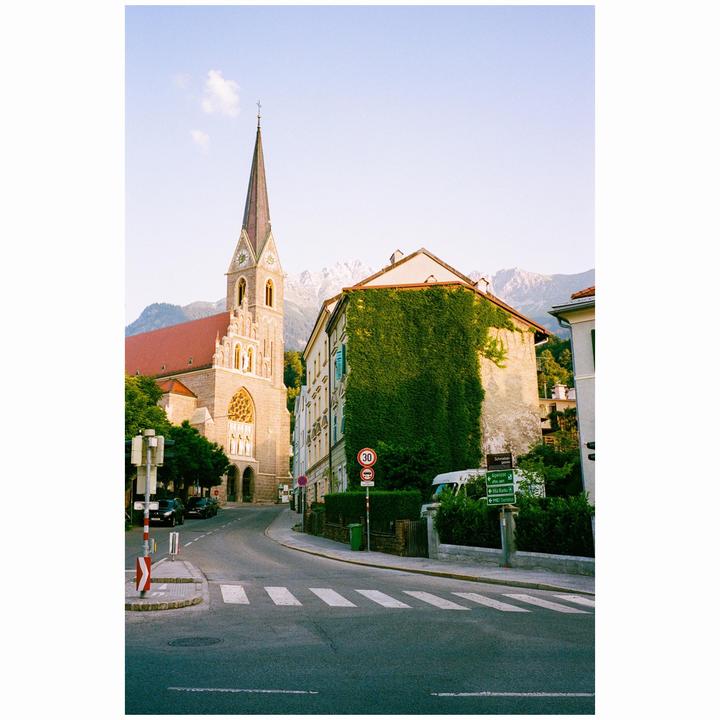 A street curves past a church with a tall spire and a building covered in ivy, with mountains in the background.