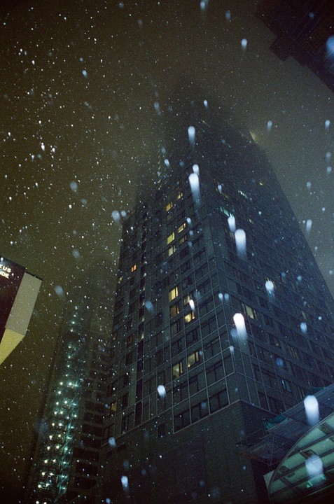 Snow falls at night in front of a tall building with many windows.