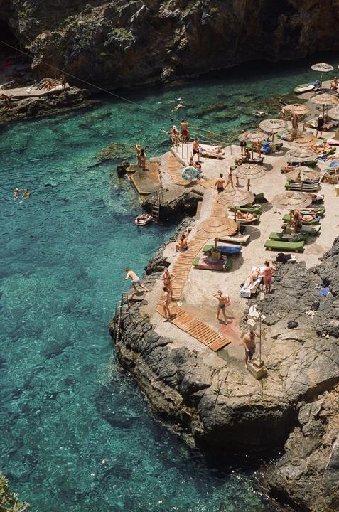 A rocky beach with many people swimming and sunbathing under straw umbrellas.