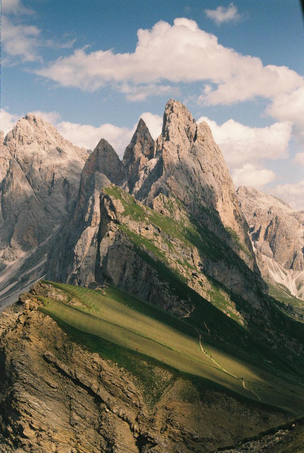 A mountain range with jagged peaks and green slopes is visible under a blue sky with white clouds.