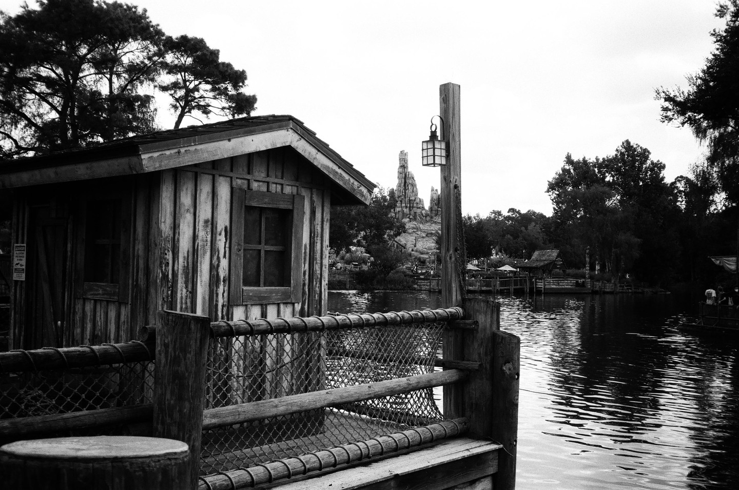 A wooden shack with a window and a fence stands next to a body of water, with a mountain in the background.