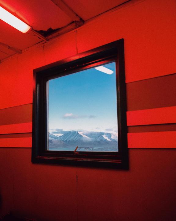 A window with a red interior frame looks out onto snow-capped mountains under a blue sky.