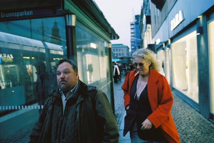 A man and a woman walk on a city street past a bus stop and storefronts.