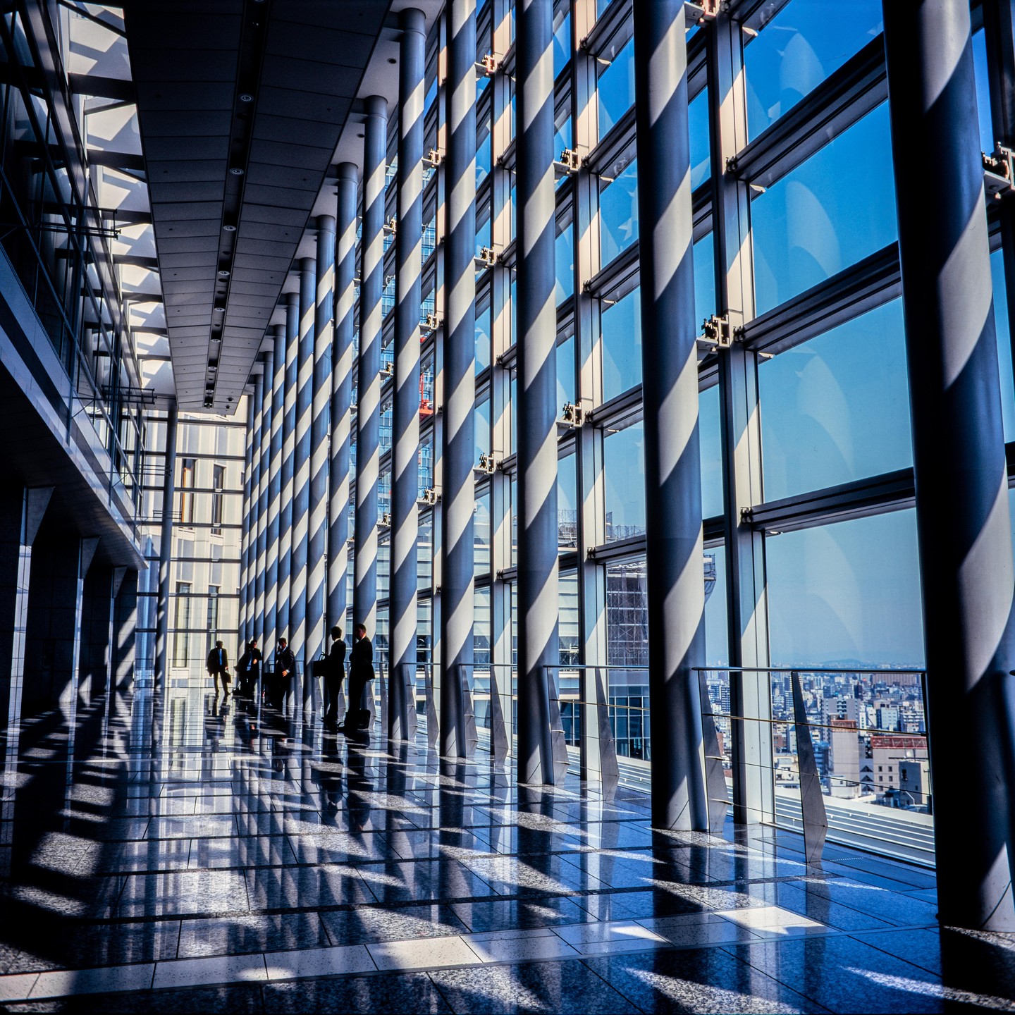 A group of people stand in a modern building hallway with tall windows and many pillars casting long shadows on the polished floor.