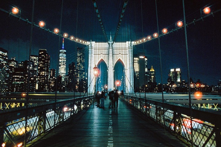 People walk across the Brooklyn Bridge at night with the New York City skyline in the background.