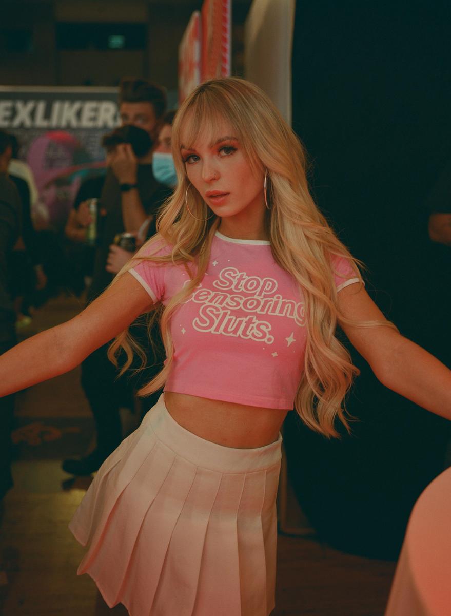 A woman with long blonde hair and bangs wears a pink crop top and a light pink pleated skirt.