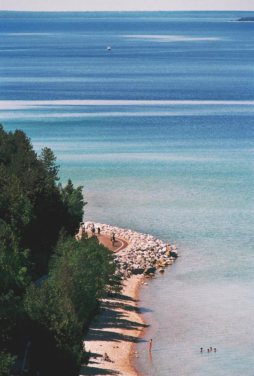 A high-angle view shows a winding path along a rocky and sandy shore with people swimming and walking, and a boat in the distance on the blue water.