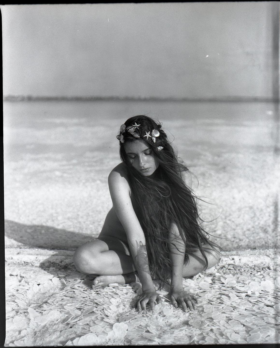 A woman with long hair and a seashell crown kneels on a salt flat.