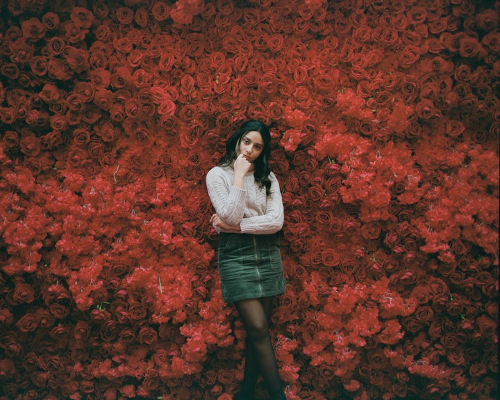 A woman stands in front of a wall of red roses and red flowers.